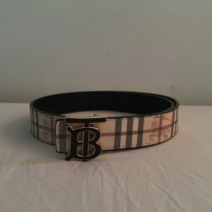 Rutig bälte  - Snyggt bälte med klassiskt rutigt mönster i beige, brun och svart. Bältet har ett brett band i skinn och ett stilrent guldspänne med BB-ikoniska logga framtill. Perfekt för att lyfta din outfit med en lyxig touch.