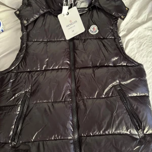 Svart dunväst från Moncler - Svart, glansig dunväst från Moncler med quiltad design och två fickor med dragkedja. Västen har en hög krage, tryckknappar vid axlarna och Moncler-logga på bröstet. Perfekt för lager-på-lager och streetstyle.