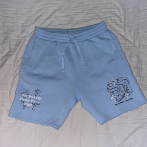 Blå shorts Supply & Demand NYC - Snygga ljusblå shorts från Supply & Demand New York City med coolt tryck och grafiska detaljer på båda benen. De har elastisk midja med snörning och är gjorda i mjuk bomull, perfekta för chill dagar. Fickor på sidorna för extra funktion.