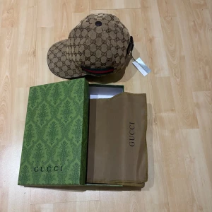 Gucci keps med GG-mönster och box - Exklusiv keps från Gucci i beige och brunt med klassiskt GG-monogram över hela kepsen. Kepsen har justerbar rem bak och ikoniska gröna och röda ränder på sidan. Levereras med originalbox och dustbag. Perfekt för dig som vill sticka ut med lyxig streetstyle.