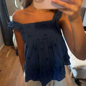 Blå offshoulder blus med volang - Supersöt blå blus med offshoulder och volangdetaljer. Toppen har broderade blommor, rynkade partier upptill och spetsdetaljer längs nederkanten. Perfekt för varma dagar och ger en romantisk vibe.