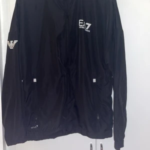 Svart vindjacka från Emporio Armani EA7 - Svart vindjacka från Emporio Armani EA7 med dragkedja framtill, huva och vita logodetaljer på bröstet och ärmen. Jackan har två fickor med dragkedja och en sportig, stilren look. Perfekt för dig som gillar streetwear och snygga märkesplagg.
