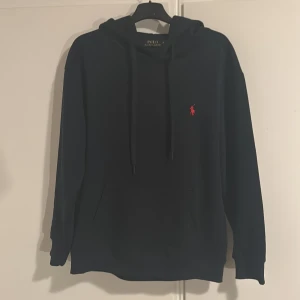 Marinblå hoodie från Polo Ralph Lauren - Svart hoodie från Polo Ralph Lauren med klassisk röd broderad logga på bröstet. Tröjan har huva med snörning, känguruficka och långa ärmar. Perfekt för dig som gillar stilren och enkel streetwear. Helt nyskick. Skriv för frågor och såklart är det äkta.