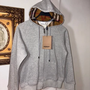 Grå Burberry hoodie med dragkedja - Snygg grå hoodie från Burberry med klassiskt rutigt foder i huvan. Tröjan har dragkedja framtill, snörning i huvan och två fickor. Perfekt för dig som vill ha en stilren och lyxig look med en touch av Burberrys ikoniska mönster.