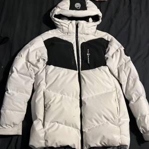 Vit och svart pufferjacka Sail Racing - Säljer en fet pufferjacka från Sail Racing i vitt med svarta detaljer. Jackan har huva, två bröstfickor med dragkedja och coola loggor på bröstet och ärmen. Perfekt för kalla dagar när du vill hålla stilen och värmen. Storlek 170 men passar exakt som S. Några små fläckar man ser på sista bilden