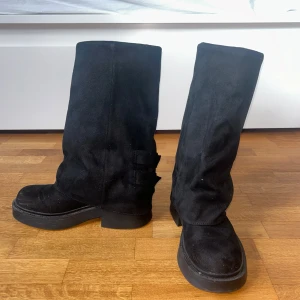 Svarta mockastövlar med spännen - Säljer ett par svarta stövlar i mocka med två snygga metallspännen på sidan. De har en platt sula och bred passform, perfekt nu till hösten/vintern!  Stövlarna är i Strl 38, har bara använts två gånger!