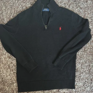 Svart stickad tröja Polo Ralph Lauren - Svart stickad tröja från Polo Ralph Lauren med röd broderad logga på bröstet. Tröjan har hög krage, half zip-dragkedja och ribbade muddar vid ärmslut och nederkant. Perfekt för dig som gillar klassisk och stilren look.