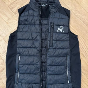 Svart dunväst från Kiwistuff - Svart quiltad dunväst från Kiwistuff med hög krage och dragkedja framtill. Västen har en bröstficka med dragkedja och logga, samt två sidofickor med reflexdetaljer. Perfekt för lager-på-lager och snygg till streetwear.