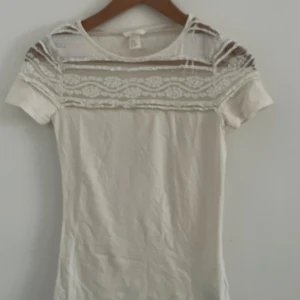 Vit blus med spetsdetaljer - Säljer en vit blus med korta ärmar och snygga spetsdetaljer upptill. Ovanför bröstet och över axlarna finns transparent spets med broderat mönster som ger en romantisk vibe. Blusen har rund halsringning och är i mjukt bomullsmaterial.