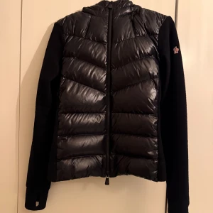 Moncler grenoble - Svart jacka med stickade ärmar, fodrad panel, klassisk huva, långa ärmar, logotypmärke på ärmen, dragkedja i fram. 