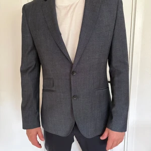 ”Mörkgråblå” kavaj från The Tailoring Club - Snygg mörkgrå blå kavaj i ren ull från The Tailoring Club. Klassisk modell med två knappar, smala slag och fickor med lock. Fodrad insida med flera innerfickor. Storlek 52. 