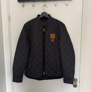 Svart quiltad jacka från Morris, S - Svart quiltad jacka från Morris med guldfärgat broderat emblem och brittisk flagga på bröstet. Jackan har dragkedja framtill, ståkrage och två fickor. Snyggt rutmönster och klassisk stil, perfekt för dig som gillar preppy vibes.