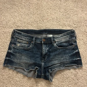 Blå jeansshorts med rå kant, H&M - Snygga blå jeansshorts från H&M med slitna detaljer och rå, fransig kant nertill. Klassisk femficksmodell med knapp och dragkedja framtill. Perfekta för varma dagar och en avslappnad look. Pris kan diskuteras!!