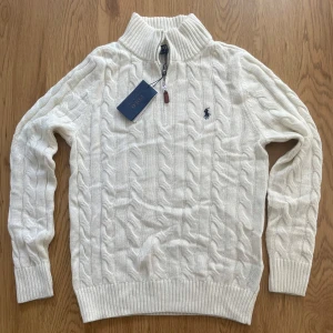 Vit kabelstickad tröja Polo Ralph Lauren - Kabelstickad vit tröja från Polo Ralph Lauren med halv dragkedja och hög krage. Klassisk broderad logga på bröstet och ribbade muddar. Perfekt för lager-på-lager och snygg till jeans eller chinos.