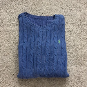 Blå kabelstickad tröja från Ralph Lauren - Kabelstickad blå tröja från Ralph Lauren med rund halsringning och klassisk grön broderad logga på bröstet. Tröjan är långärmad och har en ribbad kant vid hals, ärmslut och nederkant. Jätte fin och så fin färg!
