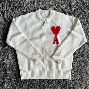 Vit stickad tröja från Ami Paris - Säljer en vit stickad tröja från Ami Paris med rött hjärta och bokstaven A broderat på bröstet. Tröjan har rund halsringning, långa ärmar och breda ribbade muddar vid ärmslut och nederkant. Perfekt för dig som gillar stilrena detaljer.
