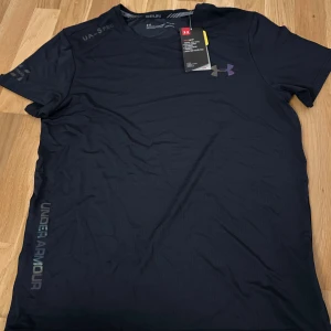 Svart tränings-tshirt från Under Armour - Svart t-shirt från Under Armour med diskret logga på bröstet och tryckt text längs sidan. Gjord i lätt och snabbtorkande polyester, perfekt för träning. Klassisk passform med rund hals och korta ärmar.