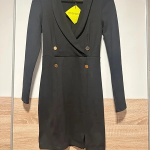 Svart kort klänning från Even&Odd - Svart kort klänning med lång ärm från Even&Odd. Klänningen har v-ringning, dekorativa guldknappar framtill och en liten slits nedtill. Snygg omlott-look upptill och figurnära passform som ger en stilren och modern vibe.