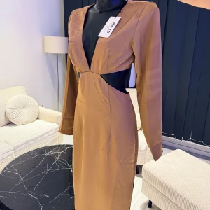 Brun långklänning med cut out från NA-KD - Snygg brun långklänning från NA-KD med djup v-ringning och cut out-detaljer i midjan. Klänningen har långa ärmar, figurnära passform och dragkedja bak. Tillverkad i ett glansigt material som ger en lyxig känsla. Passar bra i storlek 36.