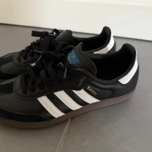 Adidas Samba svart/vit strl 40⅔ - Klassiska Adidas Samba sneakers i svart skinn med vita detaljer och bruna gummisulor. Ikoniska tre ränder på sidorna, blå logga på plösen och guldfärgad Samba-text. Snygg retrostil med snörning och rund tå, perfekt för dig som gillar streetwear.