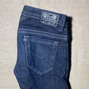 Mörkblå jeans från Diesel - Snygga mörkblå jeans från Diesel med klassisk femficksdesign och kontrastsömmar. Jeansen har en rak passform och tydlig Diesel Industry-patch baktill i grått. Tillverkade i slitstarkt denimtyg som ger en cool och tidlös look. Slim/Skinny