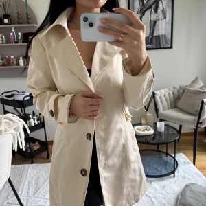 Beige trenchcoat med stora knappar - Stilren beige trenchcoat med klassisk krage och stora mörka knappar framtill. Jackan har långa ärmar, snygga detaljer vid ärmslut och midja samt två fickor. Perfekt för dig som gillar en clean och tidlös look.