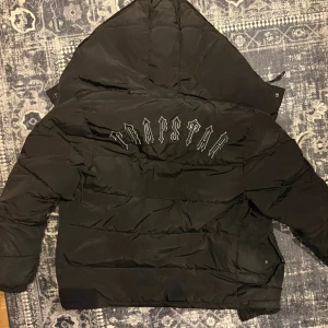 Svart pufferjacka från Trapstar - Säljer en svart pufferjacka från Trapstar med stor logga broderad på ryggen och rund patch på bröstet. Jackan har huva, dragkedja framtill och en riktigt fet, oversized vibe. Perfekt för kalla dagar när du vill se clean ut. Hål i ena fickan 