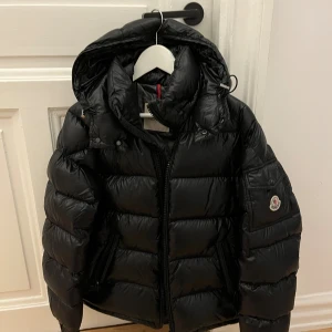 Svart Moncler pufferjacka med huva - Svart pufferjacka från Moncler, jackan har huva, dragkedja framtill och två fickor med dragkedja. Storlek 1. Köpt på NK Göteborg.  Hämtas i Vasastan, Göteborg eller fraktas via post. Jag svarar gärna på frågor eller funderingar