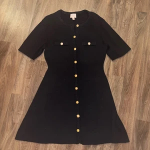 Svart kort klänning med guldknappar H&M - Svart stickad kort klänning från H&M med rund halsringning och snygga guldknappar framtill. Klänningen har korta ärmar och två dekorativa fickor på bröstet med matchande knappar. Klassisk och stilren modell med lätt utställd kjol, storlek L.