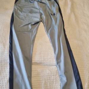 Under Armour ljusgrå träningsbyxor L - Snygga ljusgrå träningsbyxor från Under Armour med mörkblå sidopaneler och dragsko i midjan. Byxorna är tillverkade i ett mjukt och stretchigt syntetmaterial som passar perfekt för träning. Klassisk logga på låret och bekväm passform.