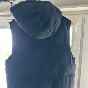 Svart dunväst med huva från Zara - Svart dunväst från Zara med stor, mysig huva och två praktiska fickor med dragkedja. Västen har en quiltad design och stängs med dragkedja framtill. Perfekt för lager-på-lager och chill stil under kyliga dagar.