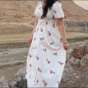 Vit långklänning med blommigt mönster - Säljer en vit långklänning med röda och gröna blommor, puffiga korta ärmar och knappar framtill. Klänningen har en feminin siluett med markerad midja och är perfekt för sommarens alla tillfällen. Materialet känns lätt och luftigt.