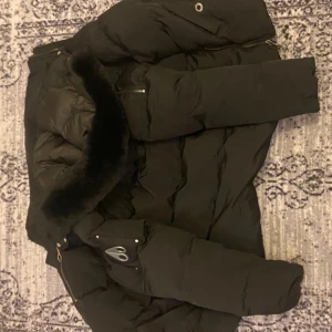 Svart dunjacka Moose Knuckles - Svart dunjacka från Moose Knuckles med fet pälskant på huvan och broderad patch på insidan. Jackan har dragkedja, långa ärmar och coola metalldetaljer på ärmen. Perfekt för kalla dagar när du vill hålla stilen.