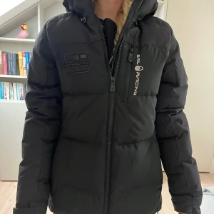 Svart dunjacka från Sail Racing - Svart dunjacka från Sail Racing med huva och logga på bröstet och ärmen. Jackan är lång, har dragkedja framtill och flera praktiska fickor. Endast använd fåtal gånger och är i väldigt bra skick. Storlek large (ungdom) men motsvarar en 36/s. Modell sail Antarctica.