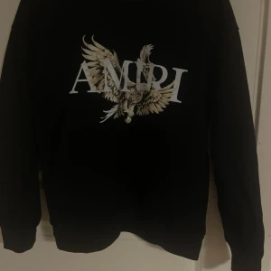 Svart Amiri sweatshirt med örntryck - Svart sweatshirt från Amiri med stort vitt logotryck och en detaljerad örn på bröstet. Tröjan har rund halsringning och långa ärmar, perfekt för dig som gillar streetwear med attityd. Säljer då jag inte använder den längre. Hör av vid frågor
