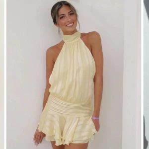 Ljusgul omlottklänning med volang  - Superfin ljusgul kortklänning med halterneck och volangkant nertill. Klänningen har subtila ränder i tyget och en bred, rynkad midja som ger en snygg siluett. Perfekt för sommarens alla tillfällen!