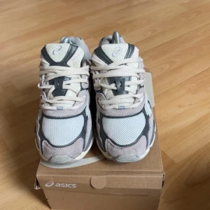 Asics sneakers i grått och vitt - Säljer ett par snygga Asics sneakers i grått, vitt och ljuslila. Skorna har mesh- och mockadetaljer, klassisk snörning och en chunky sula som ger en sportig vibe. Perfekta för dig som gillar streetstyle och bekväma skor med cool design.