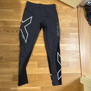 Svarta träningstights från 2XU XL - Svarta och mörkgrå träningstights från 2XU i storlek XL. Snygga reflexdetaljer och logga på benen, designade för att sitta tight och ge stöd vid träning. Tillverkade i stretchigt syntetmaterial som andas och passar perfekt för löpning eller gym.