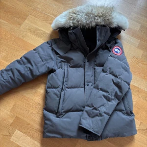 Canada Goose Wyndham - Säljer en grå dunjacka från Canada Goose med stor fluffig päls på huvan och klassisk logga på ärmen. Jackan har dragkedja och knappar framtill, två stora fickor och quiltad design. Perfekt för kalla vinterdagar och riktigt snygg. 