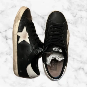 Golden Goose superstars  - Hejsan! Nu säljer vi dessa svarta Golden Goose superstars i storlek 44, skick 7/10. Skorna är i svart skinn med vit stjärndetalj på sidan och vit häl. Skorna har svarta skosnören, vit sula och GGDB-logga på plösen. Klassisk låg modell med rund tå och platt sula. Perfekta för en cool och avslappnad stil. Inget og tillkommer, pris kan diskuteras vid snabb affär och vid fler frågor är det bara att skriva! 