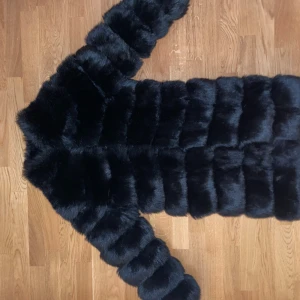 Svart lång pälsjacka i fuskpäls - Säljer en svart lång pälsjacka i fuskpäls med breda, fluffiga paneler och långärmad design. Jackan har en oversized look och är perfekt för att hålla värmen under kalla dagar. Supermjuk och ger en lyxig känsla till din outfit.