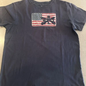 Svart t-shirt Polo Jeans Company S - Svart t-shirt från Polo Jeans Company med amerikansk flagga och broderade detaljer på bröstet. Klassisk rund hals och korta ärmar. Tillverkad i 100% bomull för en skön och avslappnad känsla. NYPRIS RUNT 3000KR