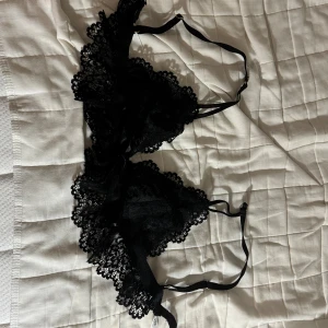 Svart spetsig bralette från Gina Tricot - Supersnygg svart bralette med spetsdetaljer och smala axelband från Gina Tricot. Toppen har en dekorativ rosett framtill och är helt i spets med justerbara band. Perfekt för att ge outfiten en extra touch.