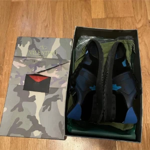 Valentino Garavani Rockrunner sneakers - Snygga Valentino Garavani Rockrunner sneakers i svart och blått med coolt kamouflagemönster. Skorna har snörning, platt sula med nitar och detaljer i skinn och textil. Perfekta för dig som vill sticka ut med exklusiva och trendiga sneakers. Pris kan diskuteras 