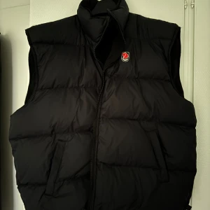 Svart Fjällräven väst - Svart/mörblå väst från Fjällräven som går att vända in och ut för att skapa två olika västar. Ena är en dunväst med puffad design, hög krage och dragkedja framtill. Den andra är en utilityväst med flera fickor, dragkedja och Fjällräven-logga på bröstet. 