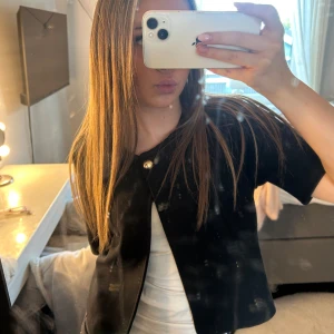 Svart kortärmad kofta med guldfärgad knapp - NY Säljer en svart kortärmad kofta med en snygg guldfärgad knapp framtill. Koftan har en rak och loose passform som ger en chill vibe. Perfekt att slänga över en topp för en enkel men stilren look.