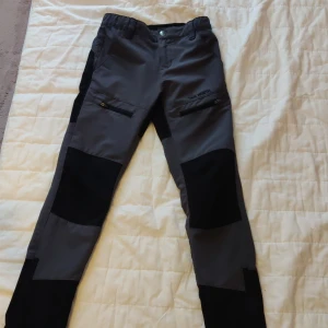 Svarta och grå täckbyxor True North 134/140 till barn - Grymma täckbyxor från True North i svart och grått. Byxorna har förstärkta partier på knän och rumpa, dragkedjor och är perfekta för uteliv. Snygg och funktionell design med elastisk midja och bälteshällor.