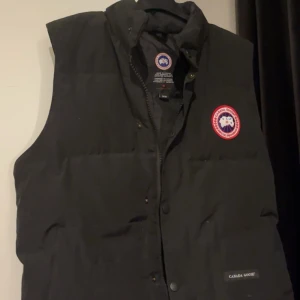 Svart dunväst från Canada Goose - Svart dunväst från Canada Goose med klassisk logga på bröstet och hög krage. Västen har två stora fickor framtill, dragkedja och tryckknappar. Perfekt för lager-på-lager och riktigt snygg till streetwear eller outdoor-stil.
