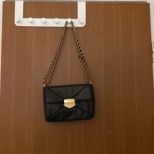 Svart axelväska med guldkedja - Snygg svart axelväska i skinnimitation med guldfärgad kedja och spänne framtill. Väskan har quiltade detaljer, ett fack med dragkedja på baksidan och flera innerfack. Perfekt storlek för det viktigaste och ger en lyxig känsla till din outfit.