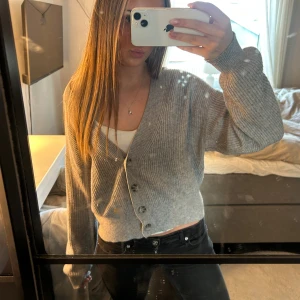 Grå ribbad kofta med knappar - Säljer en grå, croppad kofta med ribbad struktur och v-ringning. Koftan har långa ärmar och tre knappar framtill. Perfekt att slänga över en topp för en chill look. Materialet känns mjukt och är stickat, vilket gör den skön att bära.